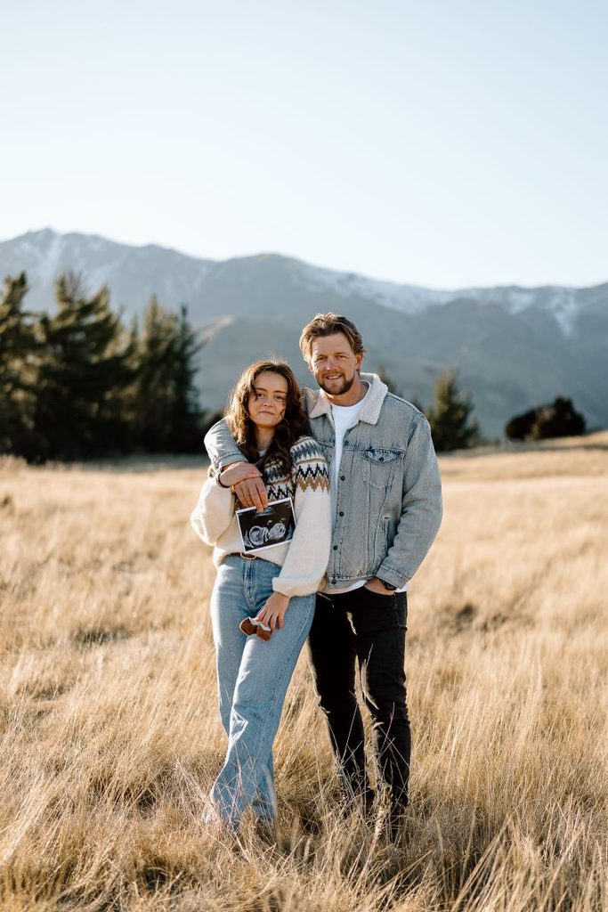 Expecting couple in the field surrounded by snowy mountains during their pregnancy announcement photoshoot at Lake Hawea, New Zealand.