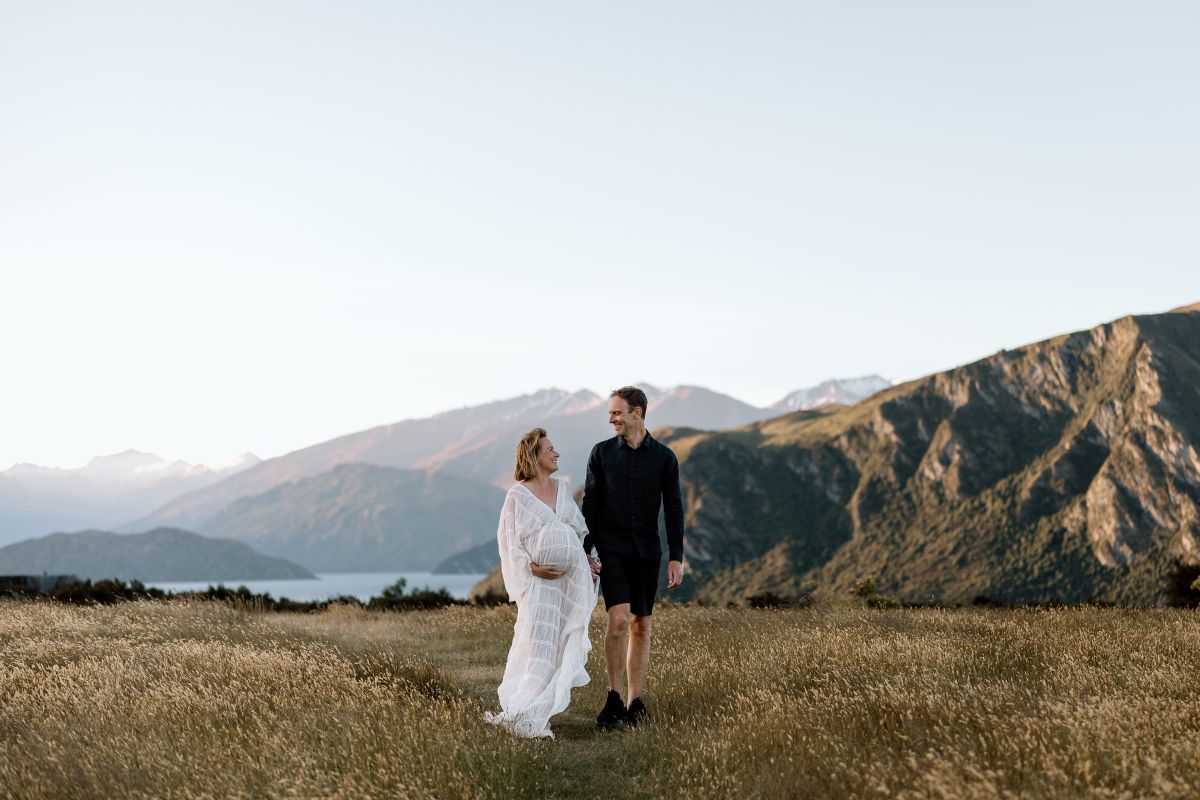 couple walking in the field during maternity photoshoot in Wanaka at Jason's Clearing with mountains and lake in the background